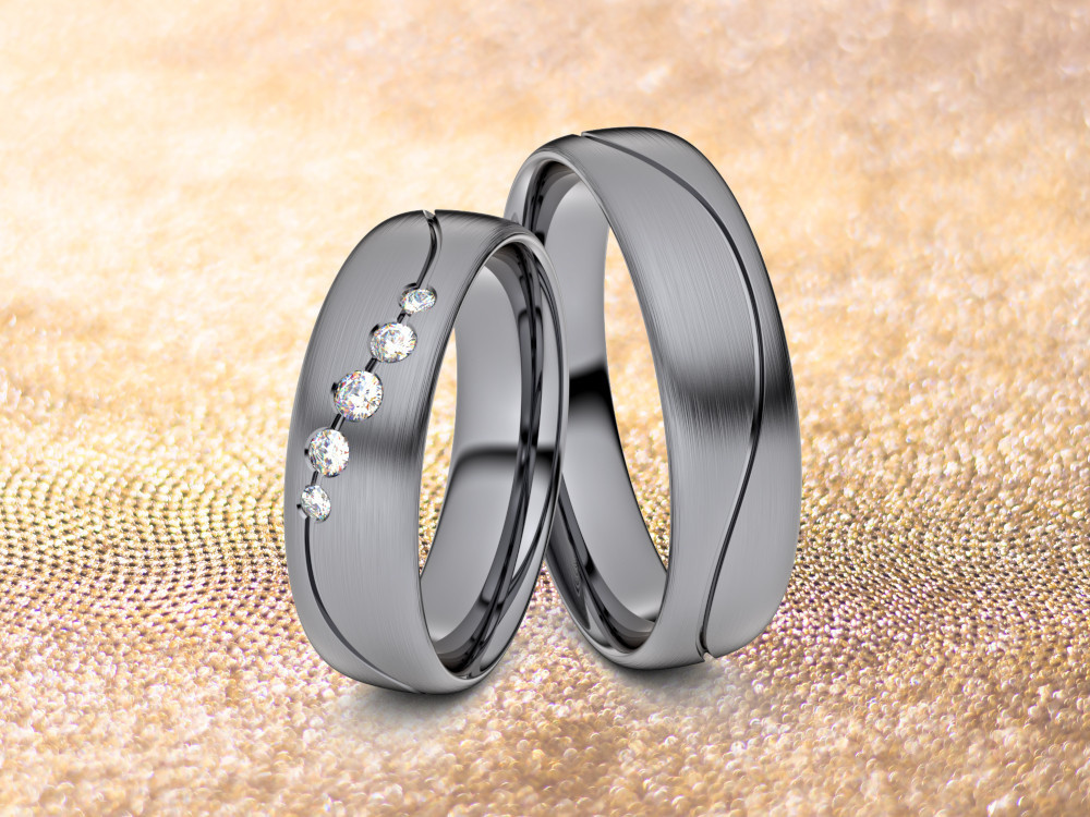 Zwei Eheringe aus Tantal sind auf einem strukturierten goldenen Hintergrund abgebildet. Ein Ring weist eine Reihe kleiner Diamanten auf, während der andere eine glatte Oberfläche hat. Die Ringe stehen aufrecht und lehnen aneinander.