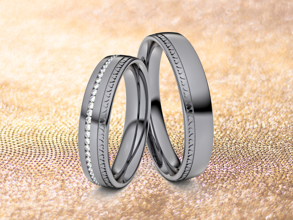 Zwei Ringe aus Tantal auf glitzerndem Goldgrund. In der Mitte des linken Rings ist eine Reihe kleiner Diamanten eingelassen, während der rechte Ring eine glatte Oberfläche mit einem dünnen gemusterten Rand hat. Beide Ringe reflektieren das umgebende Licht.