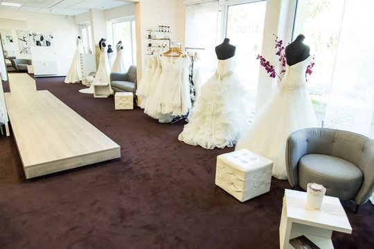 Innenansicht eines Brautmodengeschäfts mit mehreren Schaufensterpuppen in verschiedenen weißen Brautkleidern. Der Raum hat einen dunklen Teppich, helle Wände und Möbel, darunter einen grauen Stuhl und kleine weiße Tische. Auf der linken Seite befindet sich eine Ausstellungsplattform.