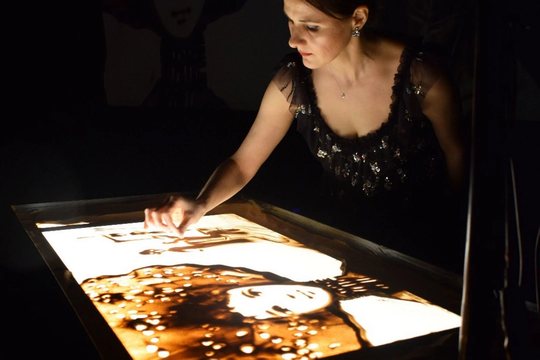 Eine Frau in einem dunklen Kleid kreiert Kunstwerke auf einem beleuchteten Sandtisch. Mit ihren Fingern formt sie den Sand zu detailreichen Bildern. Der Hintergrund ist schwach beleuchtet, wodurch das Kunstwerk hervorgehoben wird.