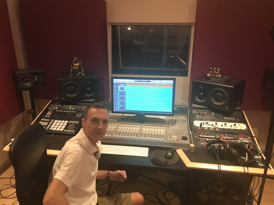 Eine Person in einem Tonstudio trägt ein weißes Hemd und beige Shorts und steht vor einem Mischpult mit Monitoren. Auf dem Bildschirm ist eine digitale Audio-Workstation zu sehen, der Raum ist mit roten Akustikplatten geschmückt.