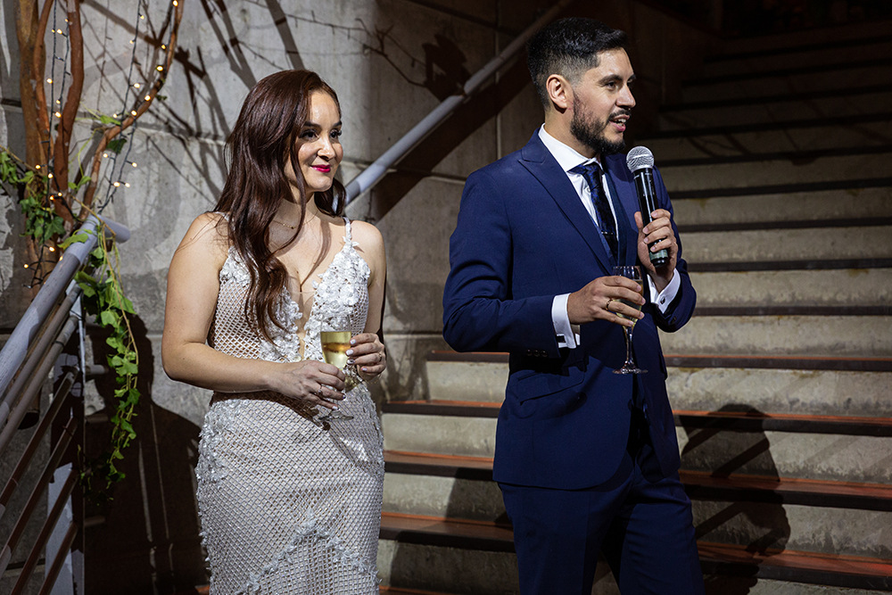 Der Bräutigam im blauen Anzug hält ein Mikrofon und ein Champagnerglas in der Hand. Neben ihm steht seine Frau im weißen Kleid, die ein Glas hält. Sie stehen an einer Treppe mit dekorativen Lichtern und Grünpflanzen.