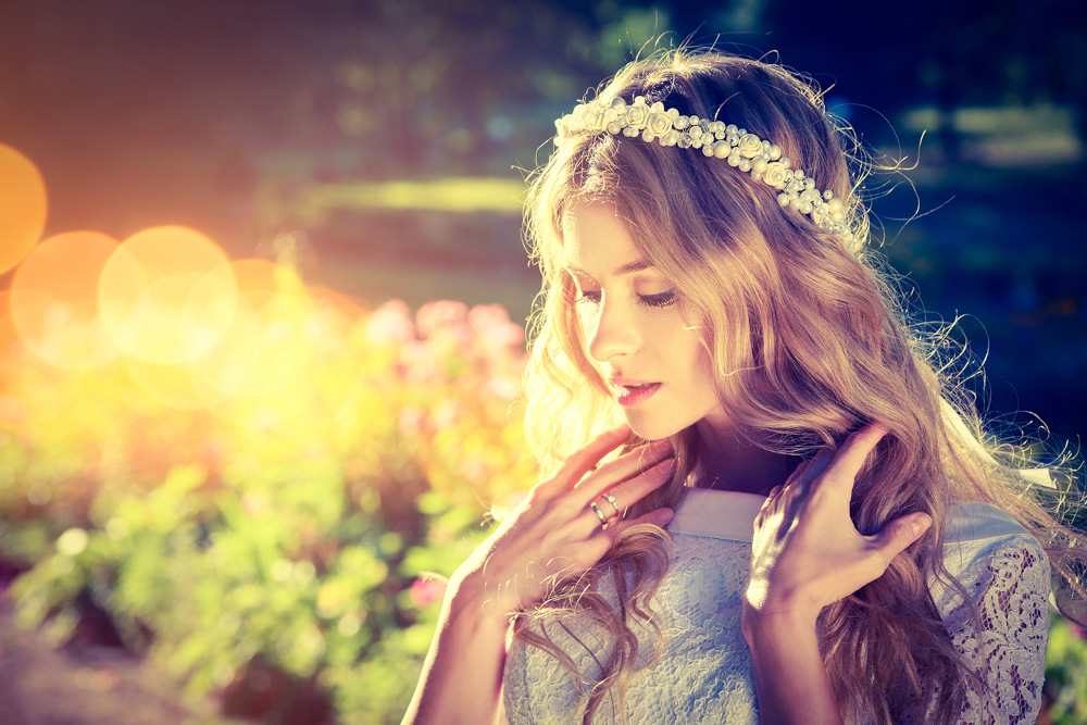 Eine Braut mit langen, welligen blonden Haaren steht im Freien. Sie trägt eine weiße Blumenkrone und ein Spitzenkleid. Sie berührt sanft ihr Haar. Ein Weichzeichnereffekt und warmes Sonnenlicht im Hintergrund schaffen eine verträumte Atmosphäre.