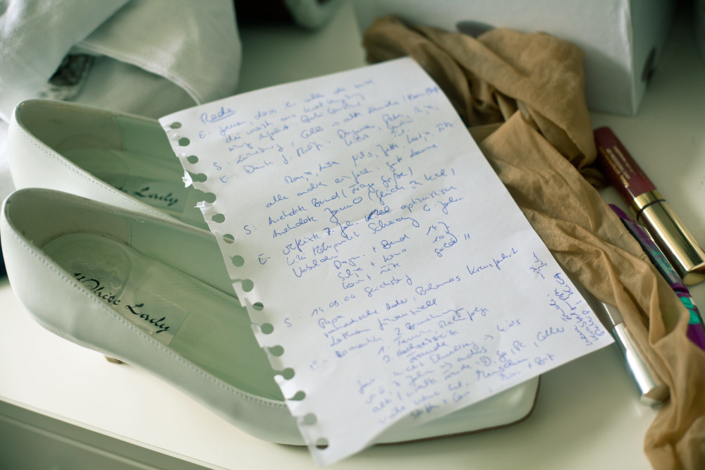 Auf einem Paar weißer Schuhe liegt eine handgeschriebene Seite mit Notizen, zu einer Hochzeitsrede. Daneben liegen ein dunkles Halstuch, ein lila Stift und ein Lippenstift.