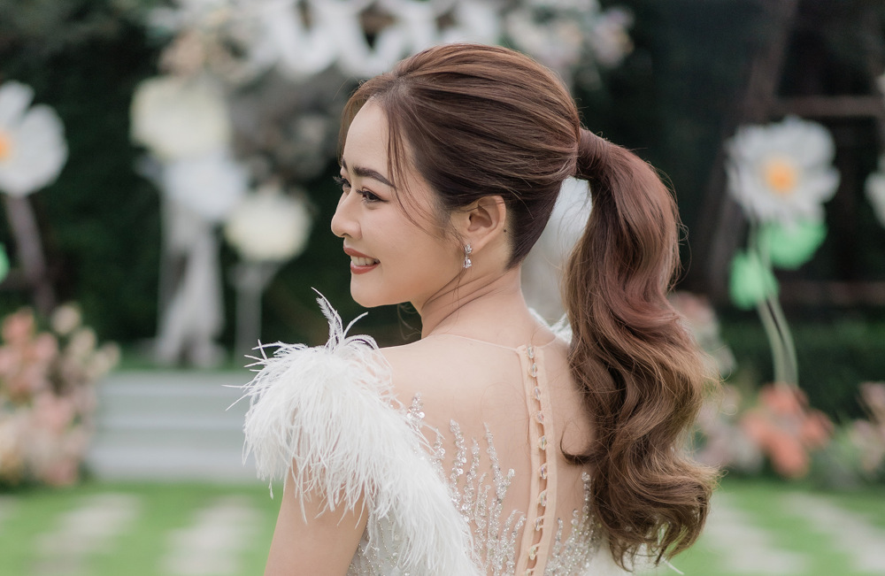 Eine Braut in einem weißen Kleid mit gefiederten Schultern und gestyltem braunem Haar lächelt, während sie in einer Außenkulisse zur Seite blickt. Der Hintergrund enthält verschwommenes Grün und große Blumen.