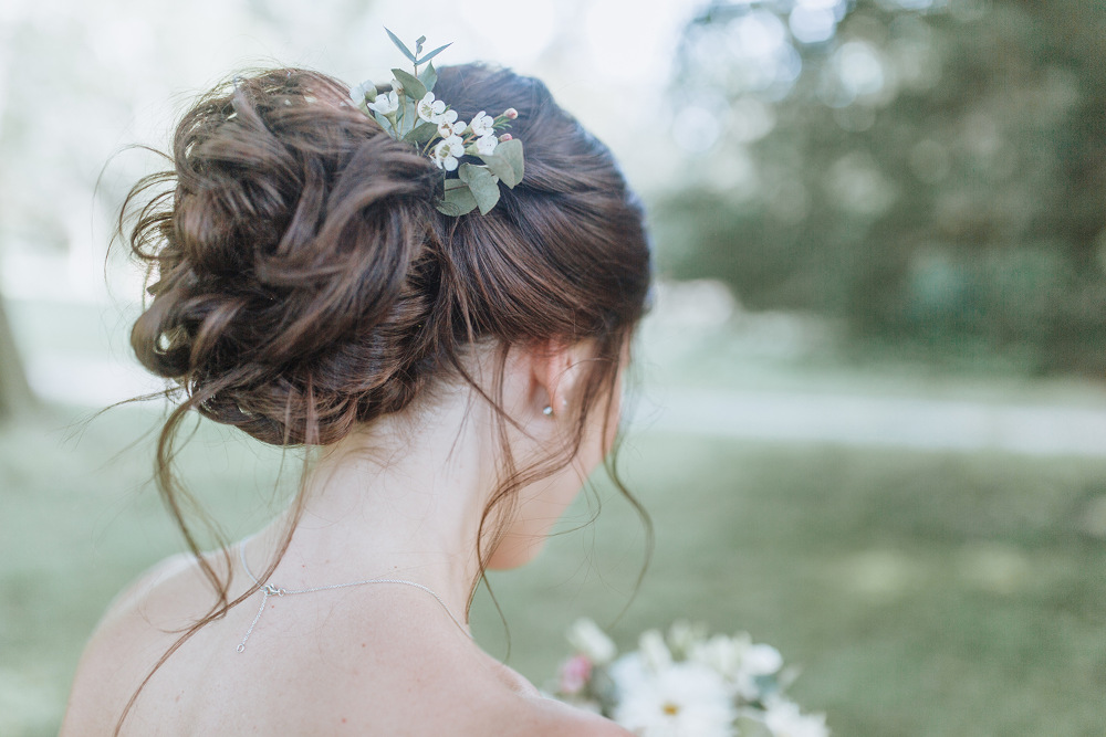 Eine Braut mit braunem Haar, das zu einer eleganten Hochsteckfrisur mit Blättern und kleinen weißen Blumen geschmückt ist, ist von hinten zu sehen. Sie hält einen Strauß weißer Blumen in der Hand und steht in einer Außenkulisse mit verschwommenem Grün im Hintergrund.