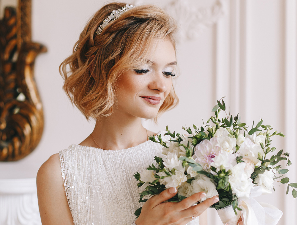 Eine Braut mit kurzen blonden Haaren und einem mit Juwelen verzierten Stirnband hält einen Strauß weißer und rosa Blumen in der Hand und betrachtet ihn. Sie trägt ein ärmelloses weißes Kleid und steht in einem Raum mit kunstvollem Dekor im Hintergrund.