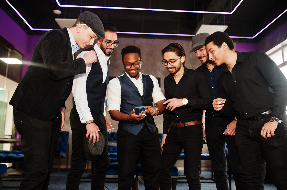 Planung des Junggesellenabschieds: Sechs Männer stehen in einem modernen Ambiente zusammen, versammelt um ein Smartphone, das einer von ihnen in der Hand hält. Sie sind leger gekleidet in Westen, Anzügen und Hüten und wirken engagiert und fröhlich, während sie auf den Bildschirm blicken.