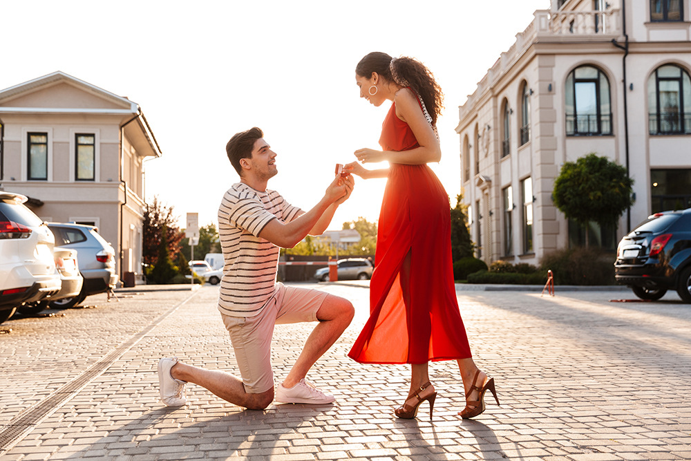 Ein Mann kniet auf einem Knie und hält einer Frau in einem roten Kleid eine Ringschachtel hin, um ihr einen Heiratsantrag zu machen. Sie sind draußen, umgeben von geparkten Autos und Gebäuden, im Hintergrund geht die Sonne unter.