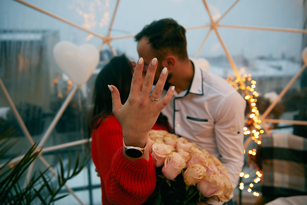 Ein Paar steht in einer geodätischen Kuppel mit Lichterketten. Die Frau trägt einen roten Pullover und zeigt einen Ring an ihrer Hand. Sie hält einen Strauß Rosen in der Hand. Der Mann im weißen Hemd steht neben ihr und hat seinen Arm um sie gelegt. Herzförmige Luftballons sind zu sehen.