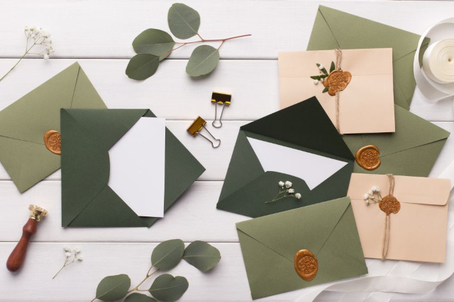 Eine Sammlung grüner und beiger Umschläge mit Wachssiegeln und Blumendekorationen auf einer weißen Holzoberfläche. Ein Wachssiegelstempel, eine goldene Büroklammer und kleine weiße Blumen sind überall verstreut.