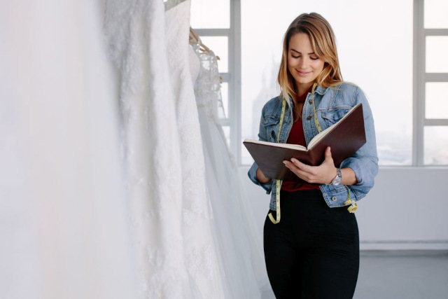 Eine Frau steht in einem hellen Raum und hält ein aufgeschlagenes Buch in der Hand. Sie trägt eine Jeansjacke und eine schwarze Hose und betrachtet das Buch mit einem leichten Lächeln. Links hängen mehrere weiße Kleider an einer Kleiderstange. Im Hintergrund sind große Fenster zu sehen.